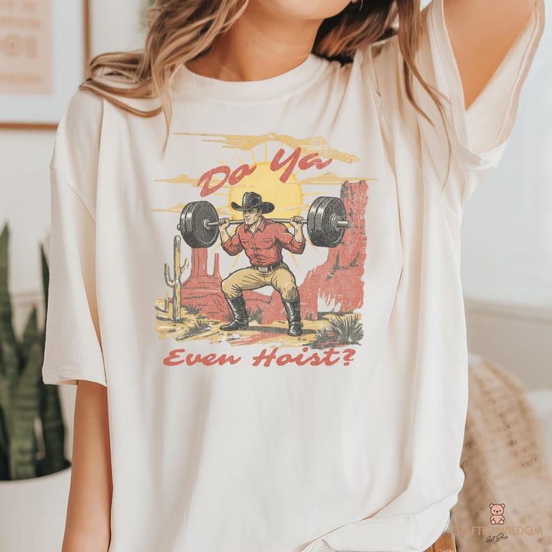 May include: Cream-colored t-shirt featuring a graphic of a cowboy lifting weights in a desert scene. The design includes the text "Do Ya Even Hoist?" in a vintage style, with a yellow sun and red rock formations.