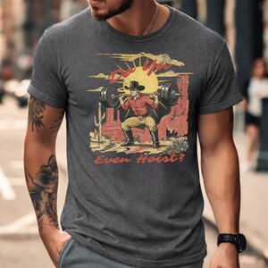 May include: Dark gray t-shirt featuring a graphic of a cowboy lifting weights in a desert scene with a setting sun. The text "Do Ya Even Hoist?" is printed on the shirt in red and yellow.