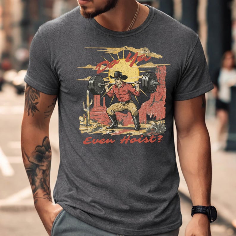 May include: Dark gray t-shirt featuring a graphic of a cowboy lifting weights in a desert scene with a setting sun. The text "Do Ya Even Hoist?" is printed on the shirt in red and yellow.