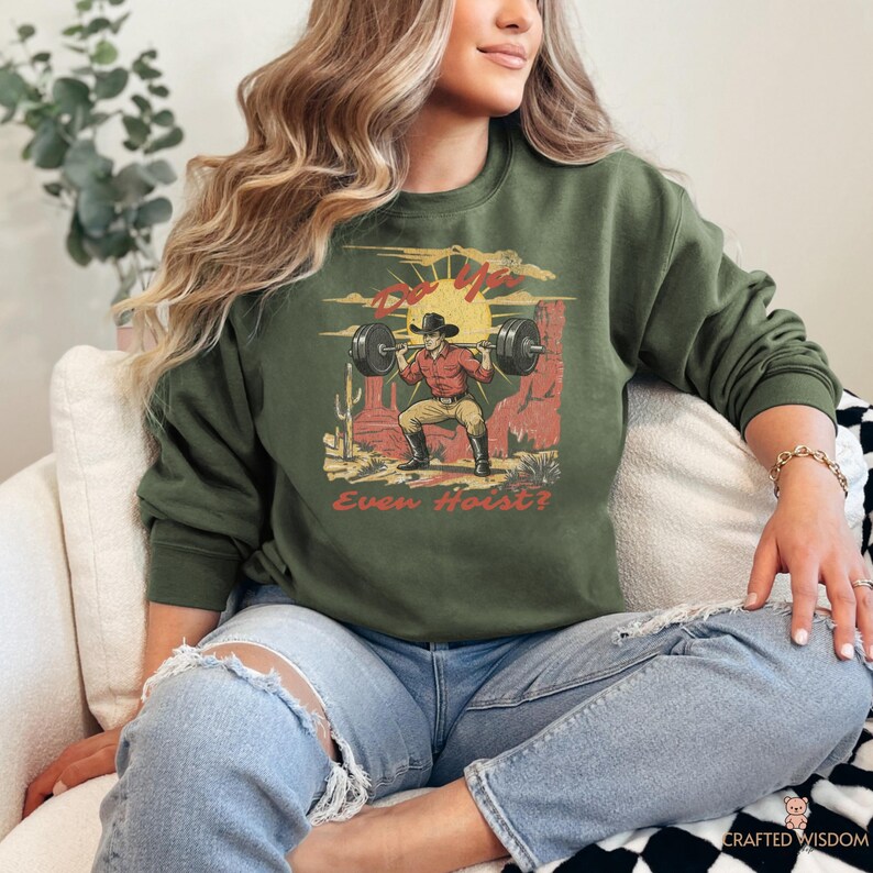 May include: Olive green sweatshirt with a graphic of a cowboy lifting weights in a desert scene. The design includes the text "Even Hoist?" in a vintage style. The cowboy is wearing a red shirt, cowboy hat, and boots.