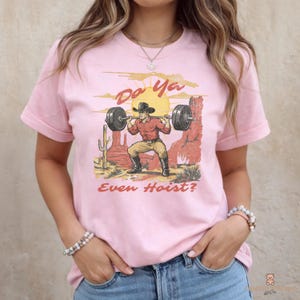 May include: Pink t-shirt with a graphic of a cowboy lifting weights in a desert scene. The text "Do Ya Even Hoist?" is printed above and below the illustration. The shirt has short sleeves and a crew neck.