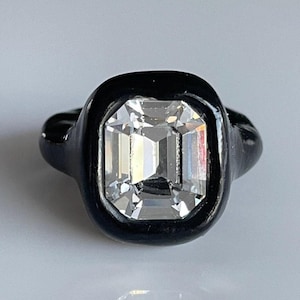 May include: A black ring with a large, clear, square-shaped gemstone set in a bezel setting.