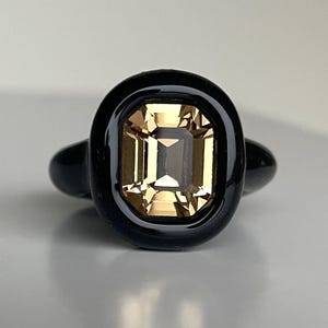May include: A black ring with a rectangular, light-brown gemstone set in a black oval frame. The ring's band is also black, creating a contrast with the gemstone's colour and shape. The ring is displayed on a light grey surface.