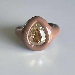 May include: A light brown gemstone ring with a pear-shaped stone. The ring has a large, clear, light brown gemstone set in a smooth, rounded, light brown band. The ring is set against a white background.