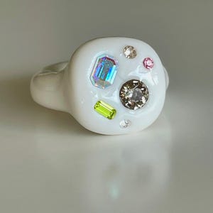 May include: A white ring with a rounded top, decorated with an assortment of colourful gemstones. The gemstones include an emerald-cut iridescent stone, a brown round stone, a pink round stone, a green rectangular stone, and several clear round stones.