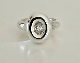 May include: A polished silver ring featuring a smooth, rounded, oval-shaped setting. At the centre of this raised silver element is a single, small, clear, oval-cut gemstone that is securely embedded within the metal surface of the ring.