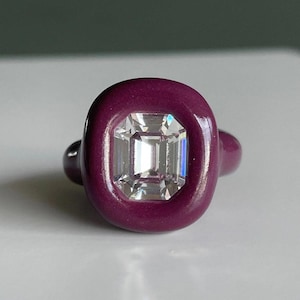 Könnte beinhalten: Ein Ring mit einer glatten, tief magentafarbenen Ringschiene und einer abgerundeten, dicken Zargenfassung. In der Mitte der Fassung befindet sich ein klarer, rechteckiger Edelstein im Smaragdschliff mit vielen Facetten.