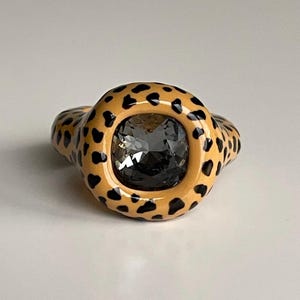 Pode incluir: Um anel com uma grande gema cinza escura engastada em uma banda espessa de cor laranja com um design de estampa de leopardo preto.