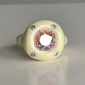 Puede incluir: Un anillo de cerámica blanca con una piedra preciosa rosa y pequeños diamantes alrededor de la piedra.