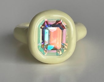 May include: A cream-coloured ring featuring a large, rectangular, iridescent gemstone. The gemstone reflects a spectrum of colours, including pink, blue, and green. The ring has a smooth, rounded band and a bezel setting.