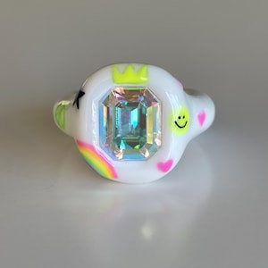 Puede incluir: Un anillo blanco con una gran gema iridiscente rectangular en el centro. El anillo está decorado con una corona amarilla, un arcoíris, un corazón rosa, una forma verde y una carita sonriente.