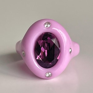 May include: A pink ring with a large purple oval-shaped gemstone surrounded by smaller clear gemstones.