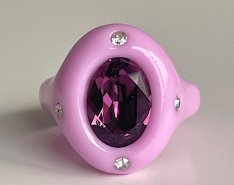 May include: A pink ring with a large purple oval-shaped gemstone surrounded by smaller clear gemstones.