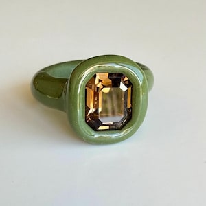 May include: A ring featuring a smooth, olive green band with a thick, rounded bezel setting. The center of the ring holds a large, rectangular, emerald-cut gemstone in a warm, translucent amber brown color with visible facets that reflect light.