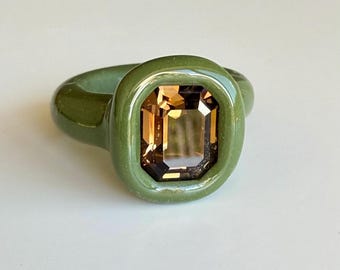 May include: A ring featuring a smooth, olive green band with a thick, rounded bezel setting. The centre of the ring holds a large, rectangular, emerald-cut gemstone in a warm, translucent amber brown colour with visible facets that reflect light.