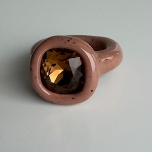 Puede incluir: Primer plano de un anillo con una gema cuadrada de color ámbar engastada en una banda rosa claro. El anillo tiene un diseño grueso y redondeado con un acabado liso y brillante. La gema refleja la luz. Una pieza de joyería.