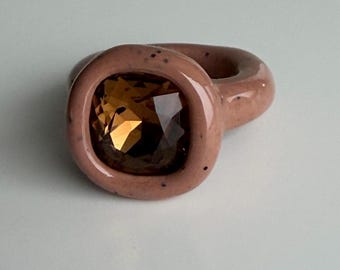 May include: A close-up of a ring featuring a square, amber-coloured gemstone set in a light pink band. The ring has a chunky, rounded design with a smooth, glossy finish. The gemstone reflects light. A statement piece of jewellery.