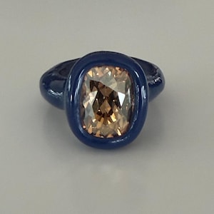 May include: A blue ring with a large, rectangular, light-brown gemstone. The gemstone is set in a bezel setting, and the ring band is also blue. The ring is a statement piece, suitable for special occasions.
