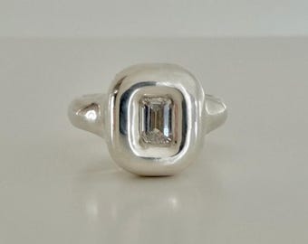 May include: A silver ring featuring a smooth, rounded, rectangular bezel setting with a polished finish. A single, clear, emerald-cut gemstone is set flush into the center of the silver frame, creating a clean and reflective appearance.