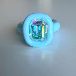 以下が含まれることがあります： 大きな長方形の虹色の宝石が付いた水色の指輪。宝石は緑、青、紫など、複数の色を反射します。指輪のバンドは、単色の水色です。