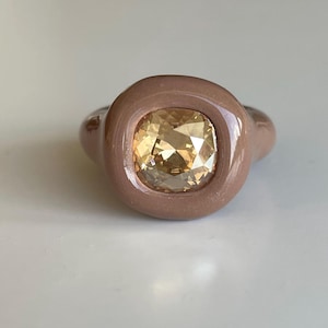 Puede incluir: Primer plano de un anillo con una gema amarilla cuadrada y facetada engastada en una banda marrón claro. El anillo tiene un diseño redondeado y liso, con un acabado pulido. La gema refleja la luz.