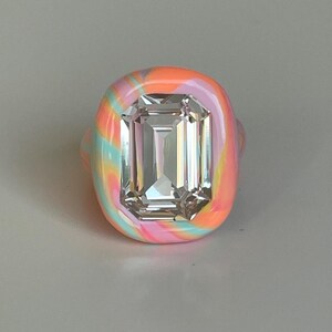 May include: A colorful ring with a large, rectangular, clear gemstone. The ring's band is a swirl of pink, orange, and turquoise. The gemstone is set in a matching, colorful bezel.