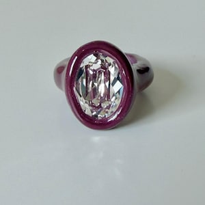 Könnte beinhalten: Ein Ring mit einem großen, klaren, oval geschliffenen Edelstein, der in ein glattes, tief pflaumenfarbenes Band gefasst ist. Die Fassung ist dick und abgerundet, was einen starken Kontrast zwischen dem dunklen Material und dem Stein erzeugt.