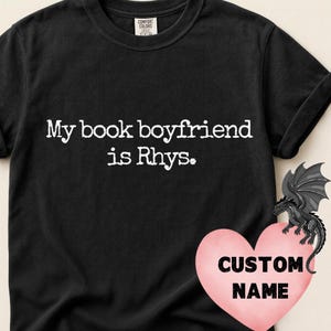 Op de afbeelding: Zwart T-shirt met de tekst "My book boyfriend is Rhys." Een roze hart met de woorden "CUSTOM NAME" en een draakafbeelding staat rechtsonder. Het shirt is gemaakt van Comfort Colors materiaal.
