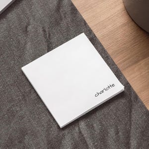 May include: A stack of white square notepads with the name "Charlotte" printed in black script. The notepads are on a dark grey fabric surface, with a wooden surface in the background.