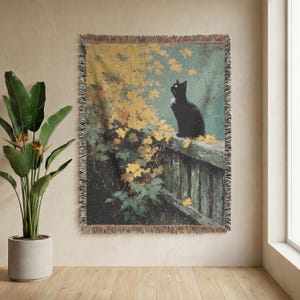 May include: A woven throw blanket featuring a black cat sitting on a stone wall. The scene includes a tree with yellow autumn leaves against a teal background. The blanket has a fringed edge and a rectangular shape with a detailed tapestry design.
