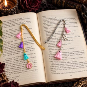 May include: Two metal hook bookmarks resting on an open book. One gold bookmark features a chain with pink, purple, and blue ghost charms and a round pink charm with the letter D. The silver bookmark has a chain with pink ghost charms and a letter R.