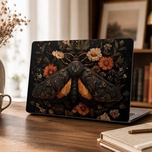 May include: A laptop skin featuring a large, dark-colored moth with orange underwings centered against a black background filled with detailed botanical illustrations of orange, white, and cream-colored flowers and green foliage.