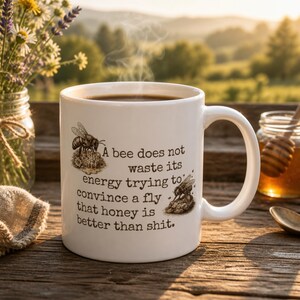 May include: White ceramic mug filled with coffee, featuring a bee illustration and the text: "A bee does not waste its energy trying to convince a fly that honey is better than shit." The mug is on a wooden surface with flowers and a jar of honey in the background.