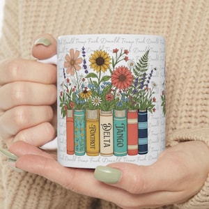 May include: White ceramic mug with a floral design, displaying colorful flowers and the words "FOXTROT", "DELTA", and "TANGO" on book spines. The mug has the phrase "Donald Trump Fuck" repeated around the rim.
