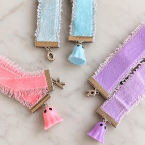May include: Four velvet ribbon chokers with frayed edges in light blue, pink, and purple. Each choker features a gold-tone metal clasp with a hanging charm, including letter charms O, K, and R, and three small ghost-shaped charms in matching colors.