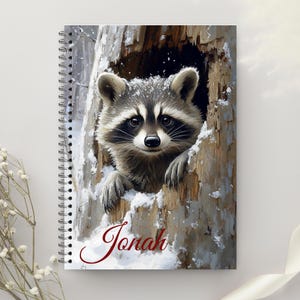 May include: Spiral-bound notebook featuring a raccoon peeking out of a snowy tree hollow. The raccoon has black and gray fur with a white face. The name "Jonah" is written in red script at the bottom. The notebook has a white background.