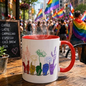 May include: White ceramic mug with a red interior and handle, featuring a colorful floral design and the word "Pride" spelled out in sign language. The mug is on a wooden table, with a chalkboard menu and a crowd with rainbow flags in the background.