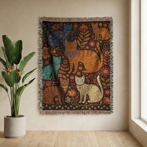 May include: A rectangular woven throw blanket featuring a dark brown background with a repeating pattern of five cats in various colors including blue, orange, teal, and cream, surrounded by stylized floral motifs and small paw prints.