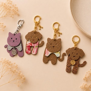 May include: Four animal-shaped keychains featuring two cats and two dogs. The first is a purple cat with a silver D charm. The second is a brown dog with a floral pattern and an A charm. The third is a brown cat with a floral bib. The fourth is a brown dog with a glittery texture and an X charm.