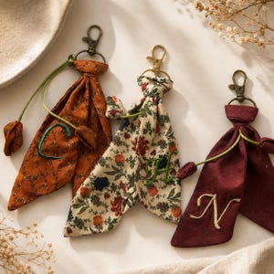 May include: Three fabric keychains with bronze-toned lobster clasps. One is burnt orange with a floral pattern and green embroidered letter C, one is cream with a colorful floral print and green letter H, and one is maroon with a gold embroidered letter N.