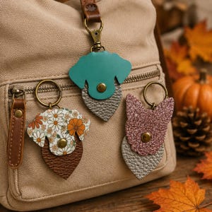 May include: Three dog-shaped keychains with metal rings and snap fasteners. One is teal with a silver textured base, one features a white and orange floral pattern with a brown base, and one is pink glitter with a silver textured base.