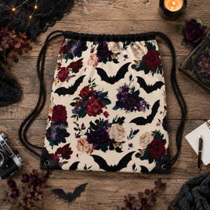 May include: A drawstring backpack featuring a cream-colored background with a repeating pattern of black bat silhouettes and clusters of deep red, dark purple, and pale pink roses. The bag includes black cord straps and black corner reinforcements.