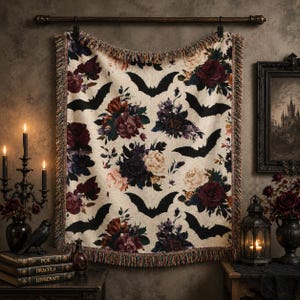 May include: A woven throw blanket featuring a cream-colored background with a repeating pattern of black bat silhouettes and clusters of deep red, dark purple, and muted pink roses. The blanket has fringed edges and is hung from a wooden rod.