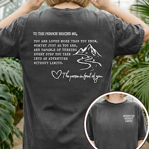 May include: Dark gray t-shirt with white text and a mountain graphic. The text on the back reads "TO THE PERSON BEHIND ME, YOU ARE LOVED MORE THAN YOU KNOW...WITHOUT LIMITS." A smaller graphic on the front says "ADVENTURE WITHOUT LIMITS."