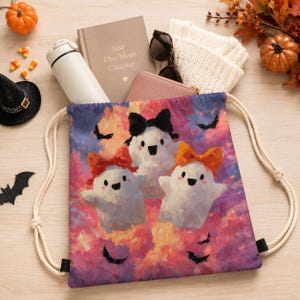 Puede incluir: Una bolsa de cordón con un estampado colorido de tres fantasmas blancos con lazos sobre un cielo rosa y morado con murciélagos negros. La bolsa tiene cordones blancos gruesos y está rodeada por una calabaza, un libro, un vaso y gafas de sol.