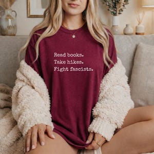 May include: A maroon t-shirt with the text "Read books. Take hikes. Fight fascists." in white. The shirt is worn with a cream-colored, fuzzy cardigan. The person is wearing gold rings and a gold necklace.