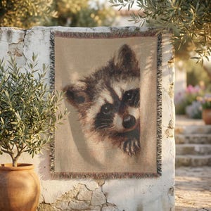 May include: A woven throw blanket featuring a detailed portrait of a raccoon with dark eye markings and a light-colored snout. The blanket has a tan background and decorative fringed edges along the top and bottom, displaying a textured weave.