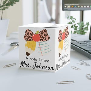 Puede incluir: Bloc de notas cúbico blanco con una ilustración colorida de un lápiz, un lazo y una manzana. El texto "A note from Mrs. Johnson" está impreso. El bloc de notas está sobre una superficie blanca.
