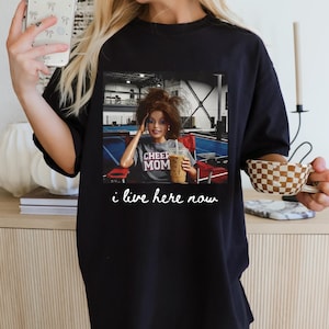 May include: Black t-shirt featuring a graphic of a doll holding a drink, with the text "CHEER MOM." Below the graphic, the text "i live here now" is printed. The shirt is worn by a person holding a phone and a cup.
