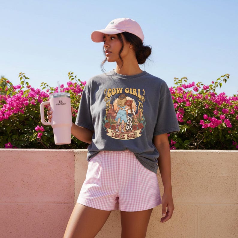 Puede incluir: Una camiseta gris carb&oacute;n de manga corta con un estampado gr&aacute;fico de dos figuras y el texto COW GIRL LIKE ME, combinada con pantalones cortos de cuadros vichy rosa claro y una gorra rosa claro, sosteniendo un vaso t&eacute;rmico grande de color rosa.
